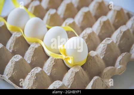 White eggs with yellow gift ribbon in an egg carton on a white background. Easter card. Stock Photo