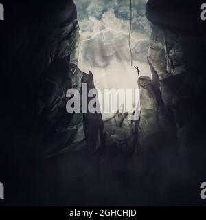 Conceptual scene with a man escaping from the steep chasm, stretching to reach the helping rope and get out of the darkness. Big cave inside the rocky Stock Photo