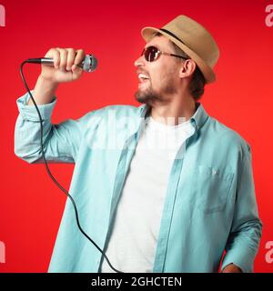 A portrait of a middle-aged man in a hat holds a microphone in his hand and emotionally sings a song. Singer in casual clothes on a red background. Stock Photo