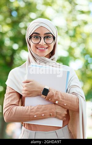 Cheerful glad cute young arab muslim female student in hijab and glasses holds books, ready for lecture Stock Photo