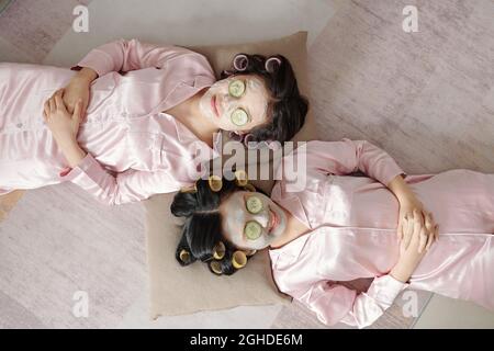 Joyful female friends relaxing with purifying clay mask and cucumber slices on face, view from above Stock Photo