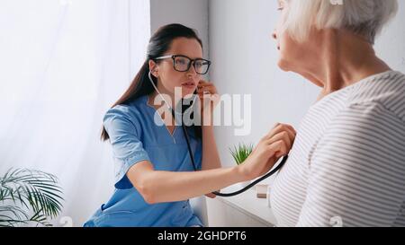 brunette nurse in eyeglasses examining senior woman with stethoscope Stock Photo