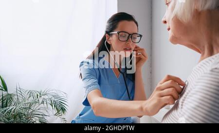 brunette nurse in eyeglasses examining aged woman with stethoscope Stock Photo