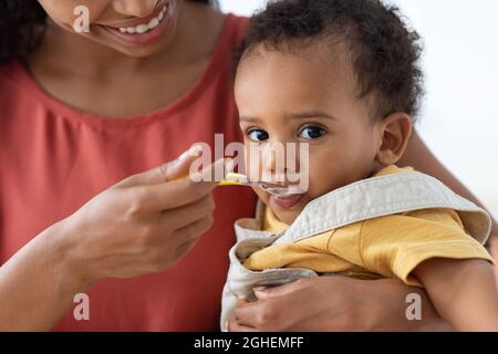 Happy Young Black Mother Spoon Feeding Her Cute Infant Baby Son, Closeup Stock Photo