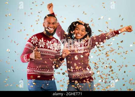 Excited young black couple in winter sweaters celebrating Christmas or New Year, having fun under confetti Stock Photo