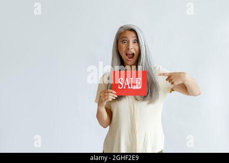 Excited mature Asian woman with silver hair points onto red Sale sign on light background Stock Photo