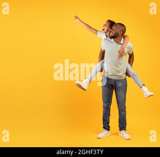 Discounts for family and kids. Excited black man giving piggyback ride for daughter, cute girl pointing at empty space Stock Photo