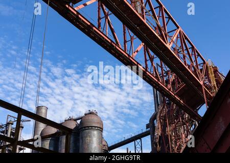 View looking up through rusting metal structures of a steel manufacturing plant, blast furnaces behind, blue sky with light clouds, horizontal aspect Stock Photo