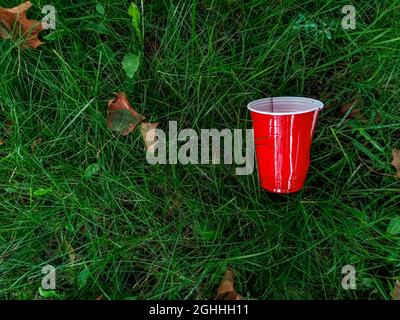 Used red plastic water coffee tea cup thrown on a grass lawn - symbol of pollution, concept Stock Photo