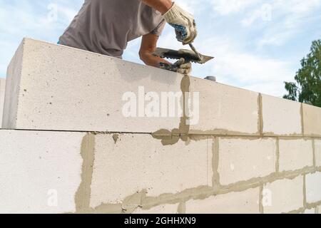 Worker wipes the seams with a spatula outside the house of blocks. Bricklayer worker laying white blocks of a future house in the open air. Stock Photo