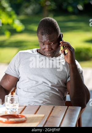 Handsome Afro American man sitting in garden at table. Attractive young man listening having phone call. Stock Photo