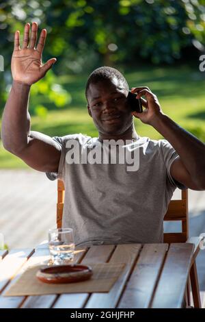 Handsome young man greeting waving at camera sitting outdoors. African man talking on phone sitting in yard smiling. Stock Photo