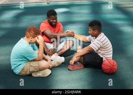 Smiling young man shaking hand of teenage boy while they sitting on ground and resting after basketball game Stock Photo