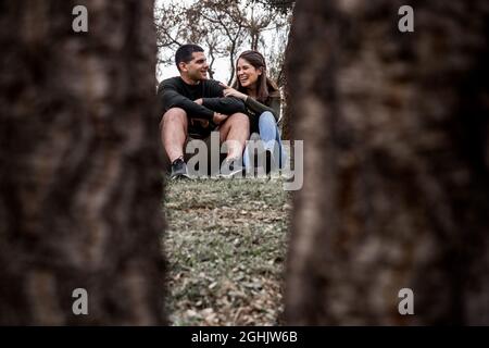Hispanic couple happily talking while sitting on grassy field of a park view through wooden trunks Stock Photo