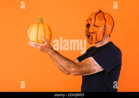 Man in profile, wearing terrifying pumpkin latex mask with a blue t-shirt, holds a 'Jack-o-lantern' pumpkin and looks at it, on an orange background. Stock Photo