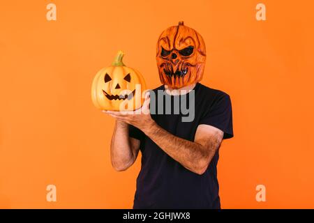Man wearing scary pumpkin latex mask with blue t-shirt, holding 'Jack-o-lantern' pumpkin on orange background. Halloween and days of the dead concept. Stock Photo