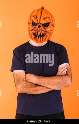 Man wearing scary pumpkin latex mask with blue t-shirt with crossed arms, on orange background. Halloween and days of the dead concept. Stock Photo
