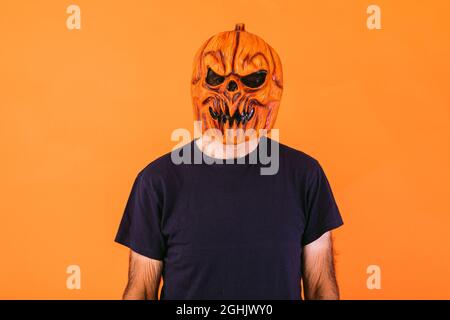 Man wearing scary pumpkin latex mask with blue t-shirt on orange background. Halloween and days of the dead concept. Stock Photo