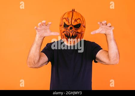 Man wearing scary pumpkin latex mask with blue t-shirt scares with his hands, on orange background. Halloween and days of the dead concept. Stock Photo
