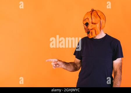 Man wearing terrifying pumpkin latex mask with blue t-shirt, pointing to the left side with his hands at the copy space, on an orange background. Hall Stock Photo