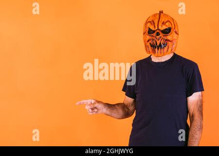 Man wearing terrifying pumpkin latex mask with blue t-shirt, pointing to the left side with his hands at the copy space, on an orange background. Hall Stock Photo