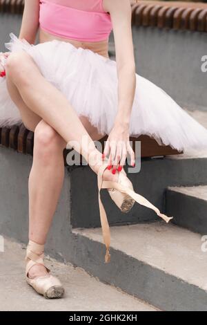 Unrecognizable Ballet dancer sits on bench and puts on beige pointe shoes. She wears light dancewear. Ballerina in city street Stock Photo