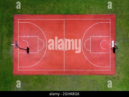 From above aerial view of empty red basketball sports field with white markings and hoops surrounded by green grass Stock Photo