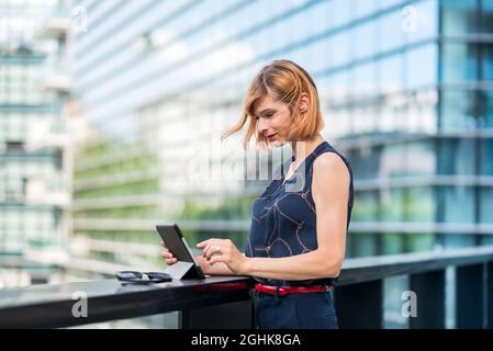 Side view of stylish young businesswoman in classy outfit using tablet while standing near railing against contemporary city building with glass walls Stock Photo