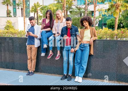 Group of diverse young men and women resting near barrier and chatting in social media on mobile phones on sunny summer day on street Stock Photo