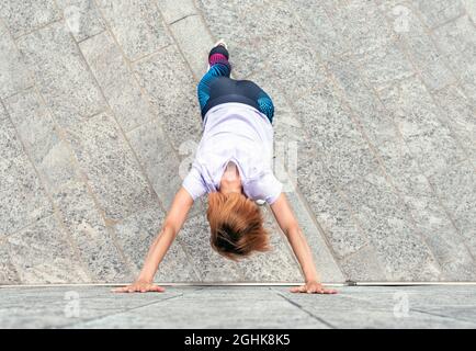 Athletic woman doing stretching exercises on a sidewalk in town in a view from above of her leaning against a building wall with arms outstretched in Stock Photo
