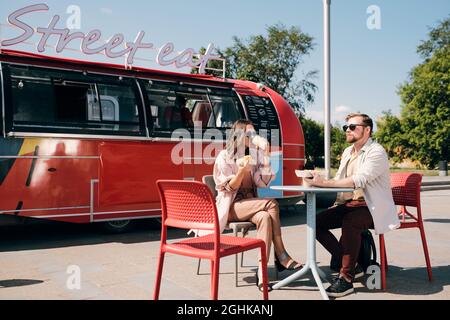 Young couple having snacks and drinks by red food truck in outdoor cafe Stock Photo