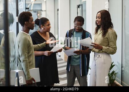 Group of young Black business people standing in corridor of modern office and discussing charts and sale reports Stock Photo