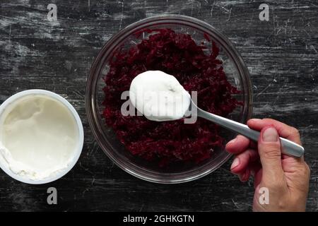 Top view of grated cooked beet in glass bowl and woman hand putting sour cream with spoon into salad on black background Stock Photo