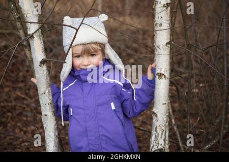 portrait of a 4 year old European boy in a gray winter fur hat and purple jacket on a walk in the forest or park. Stands between two tree trunks. Wint Stock Photo