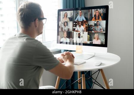 Morning meeting on the distance. Back view over shoulder of guy to the computer screen with a lot of diverse people on it, work team brainstorming, discussing together, virtual meeting colleagues Stock Photo