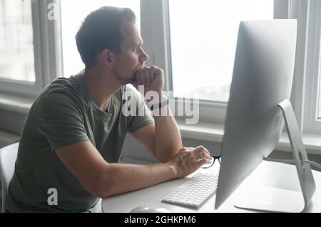 Pencive intelligent young man in casual t-shirt sits at the desk with trendy PC and looks away lost in thoughts. A freelancer takes a break and thinking about solving business tasks Stock Photo