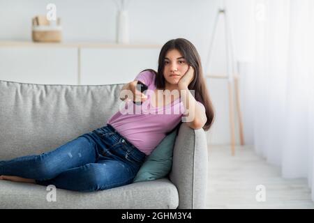 Bored Indian teen girl with remote control watching TV on sofa at home, copy space Stock Photo