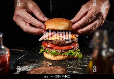 Chef preparing a gourmet double-decker beef burger with cheese, bacon rashers , salad trimmings and succulent homemade patties in a close up on his ha Stock Photo