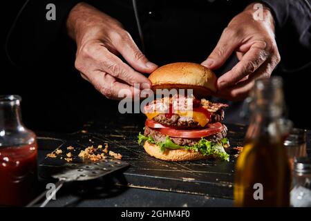 Chef or cook preparing a gourmet Whopped or double burger with succulent homemade beef patties, bacon, cheese and salad greens in a close up on his ha Stock Photo