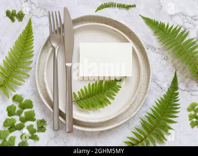Blank paper card on table setting decorated with fern leaves on white marble table top view. Tropical mock-up scene with place card flat lay Stock Photo