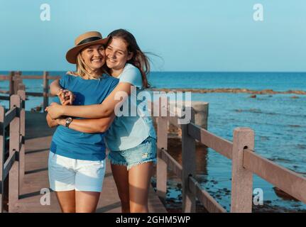 Daughter embracing Mother and standing on wooden sidewalk by the sea at sunset. A middle-aged woman and teenage girl standing side by side and wearing Stock Photo