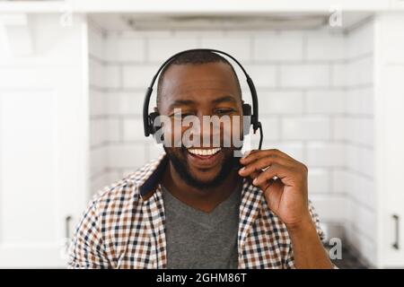 Smiling african american man sitting at countertop in kitchen making video call using headset Stock Photo