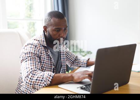 African american man sitting at table in dining room, working remotely using laptop Stock Photo