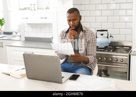 Serious african american man sitting in kitchen working looking at paperwork and using laptop Stock Photo