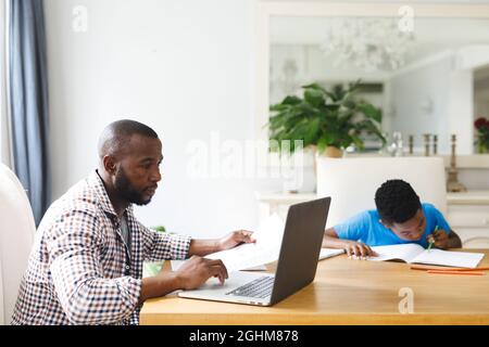 African american father working on laptop in dining room with son sitting with him doing homework Stock Photo