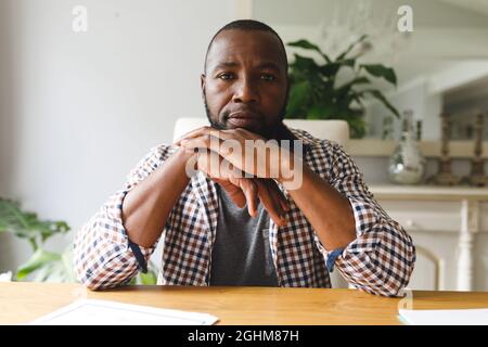 Serious african american man sitting at table in dining room, listening during video call Stock Photo