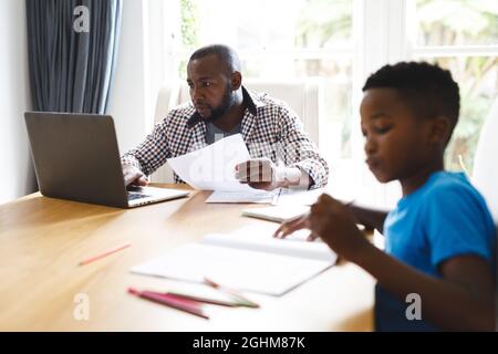 African american father working on laptop in dining room with son sitting with him doing homework Stock Photo