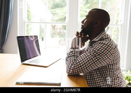 African american man sitting at table in dining room, working remotely using laptop Stock Photo