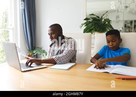 African american father working on laptop in dining room with son sitting with him doing homework Stock Photo