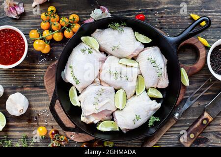Top view of a raw uncooked chicken thigh meat in a cast iron frying pan with fresh ingredients of limes, spicy sauce, lemon thyme, peppers, tomatoes a Stock Photo